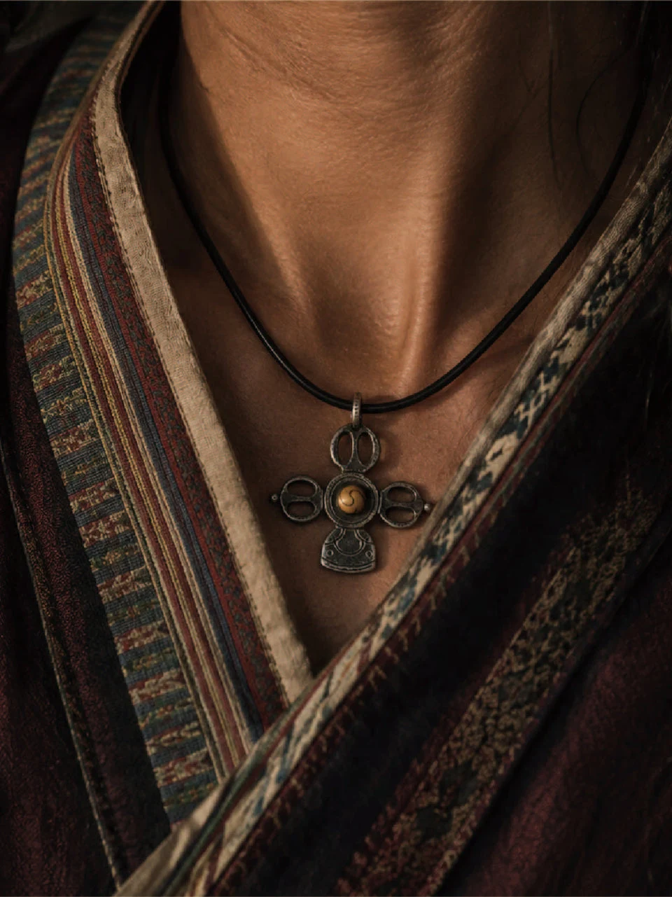 A close-up of a person wearing a black cord necklace with a dark metal pendant featuring a small gold-toned circular center, styled with patterned dark clothing.
