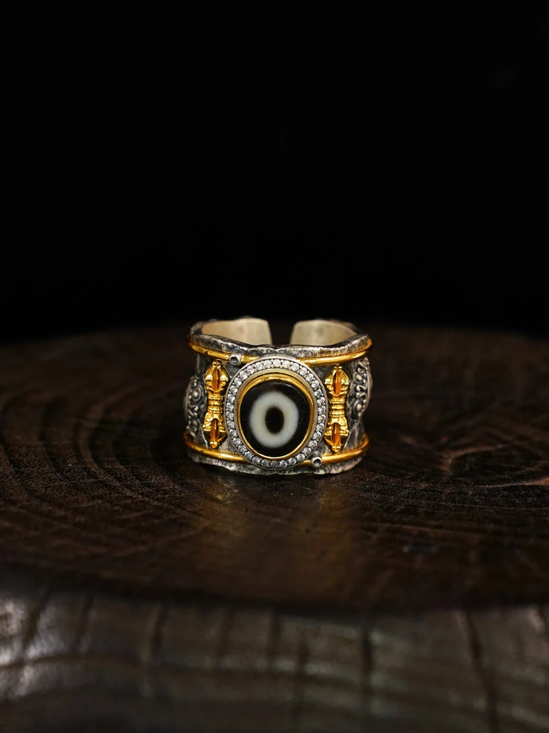 A wide silver and gold band ring with a central evil eye motif, resting on a dark wooden surface against a black background.