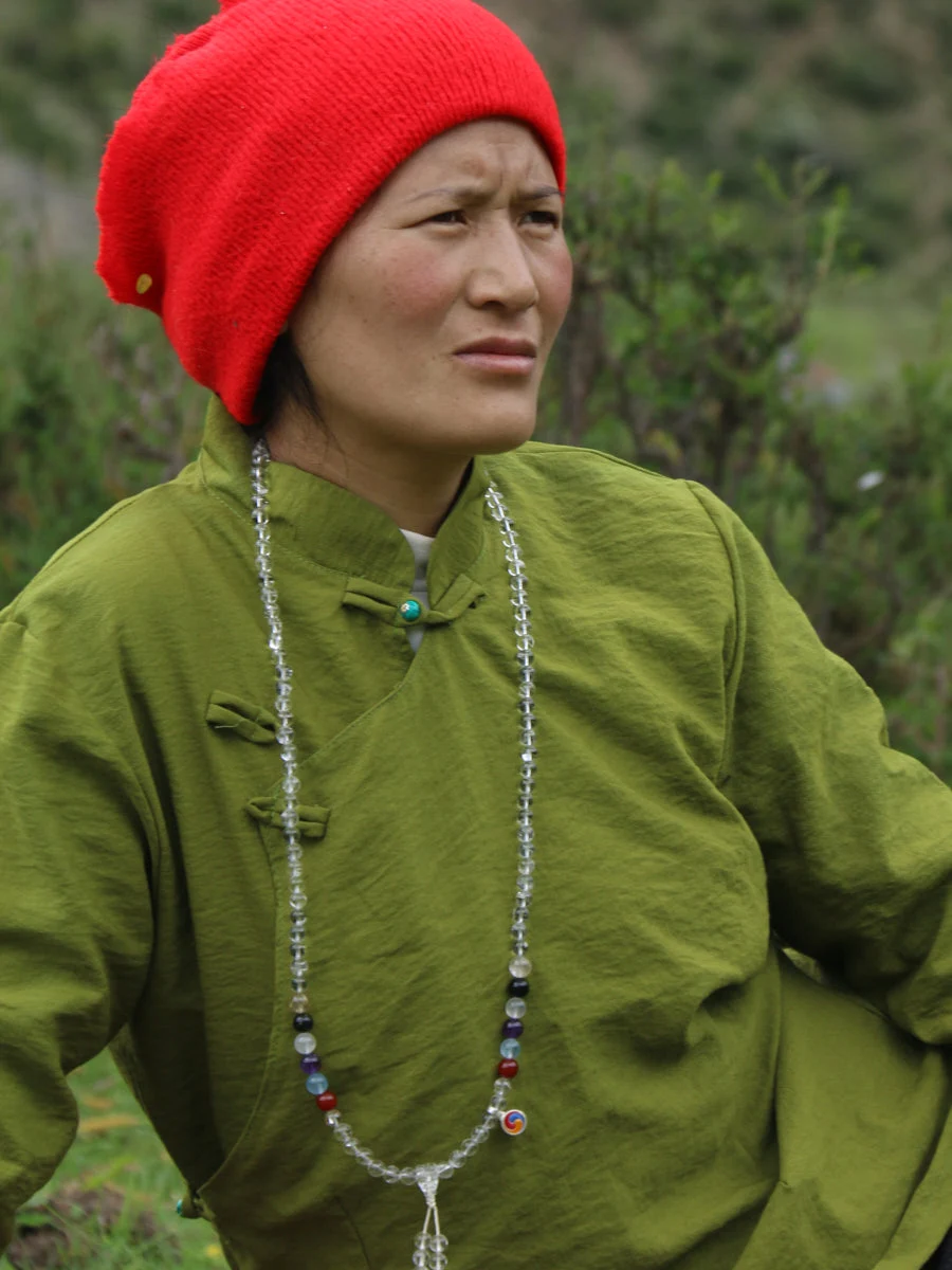 A person outdoors wearing a bright red knit beanie, an olive green traditional-style jacket, and a long beaded necklace with a small heart charm.