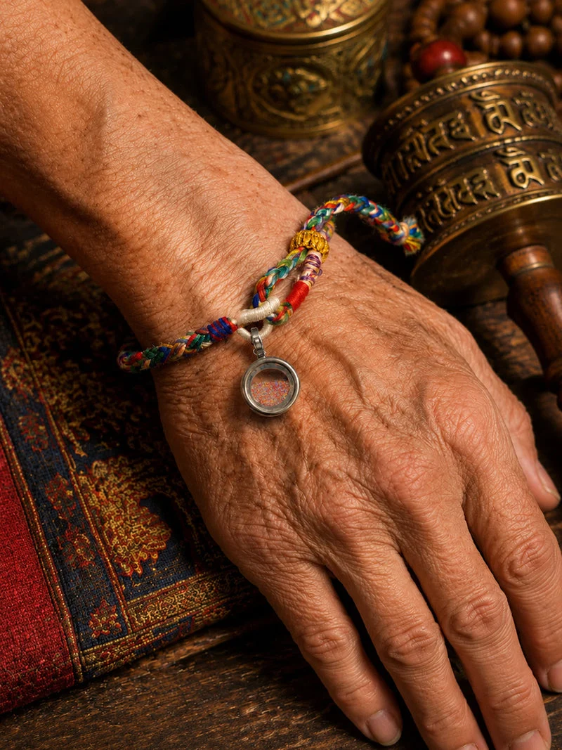 A close-up of an elderly person's weathered hand and wrist wearing a colorful braided prayer bracelet with a small circular pendant, set against a rustic background with brass prayer wheels and a patterned textile.