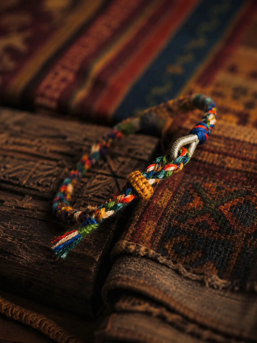 A multicolored braided cord bracelet with a white woven connector and a gold-toned accent bead, resting on a dark wooden surface with a patterned rug in the background.