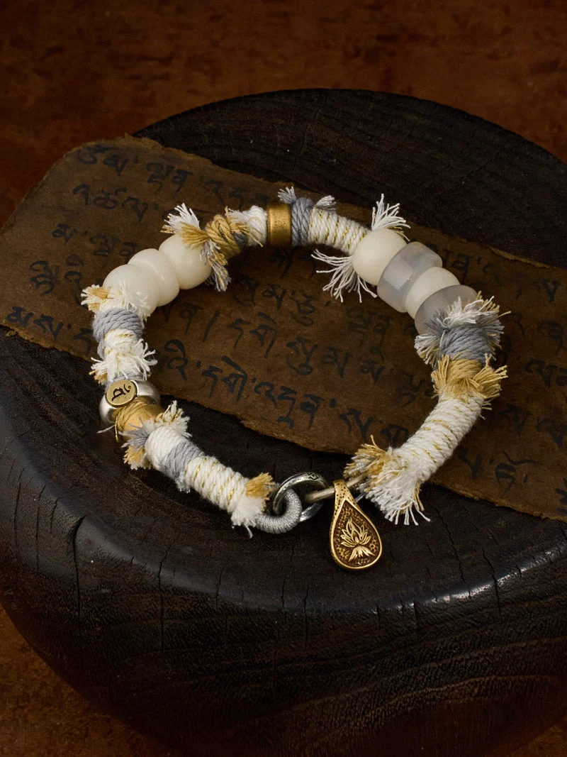 A handmade beaded bracelet with white, gray, and beige threads, gold-toned beads, and a small gold teardrop charm, displayed on a dark wooden surface with a piece of script paper.