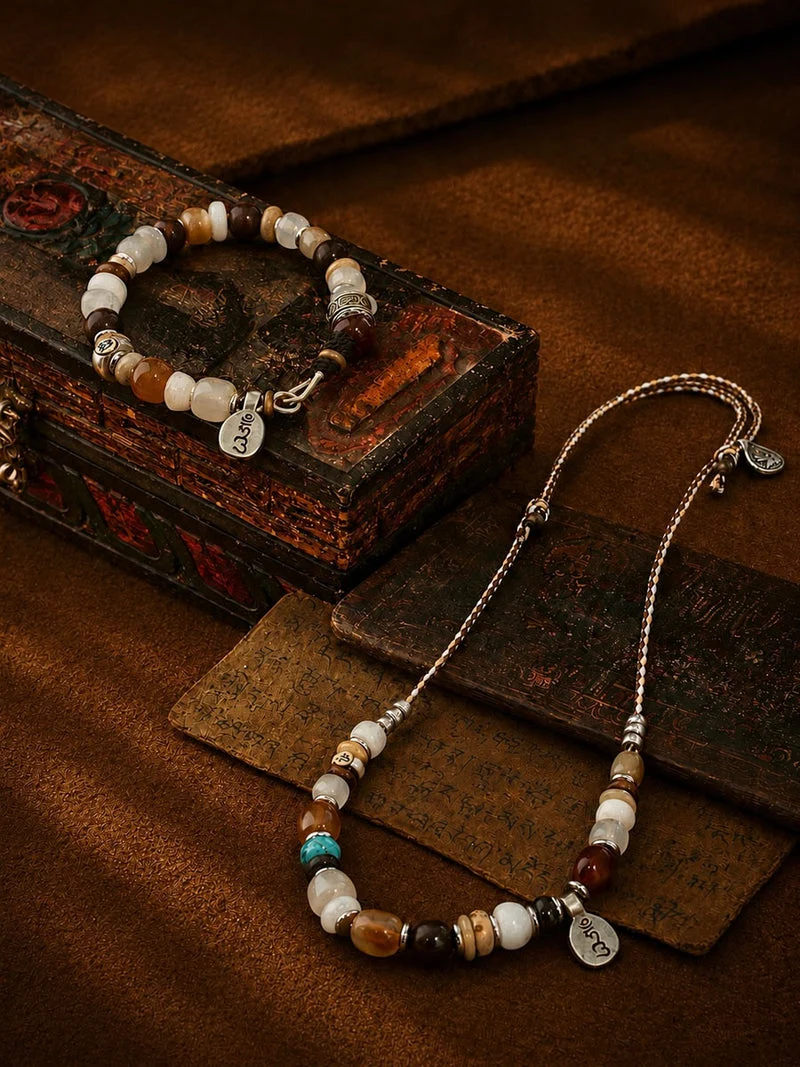 A beaded necklace and matching bracelet displayed on a rustic dark wooden box and leather surface. The jewelry features white, brown, and beige beads with small metallic spacer beads and silver-toned charms, including one engraved with 'LOVE'.