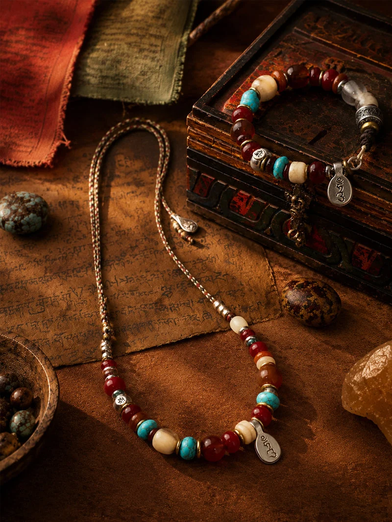A beaded necklace and matching beaded bracelet displayed on a rustic wooden surface with fabric, stones, and a small box as styled props. The jewelry features red, turquoise, cream, and metallic silver beads with engraved silver charms.