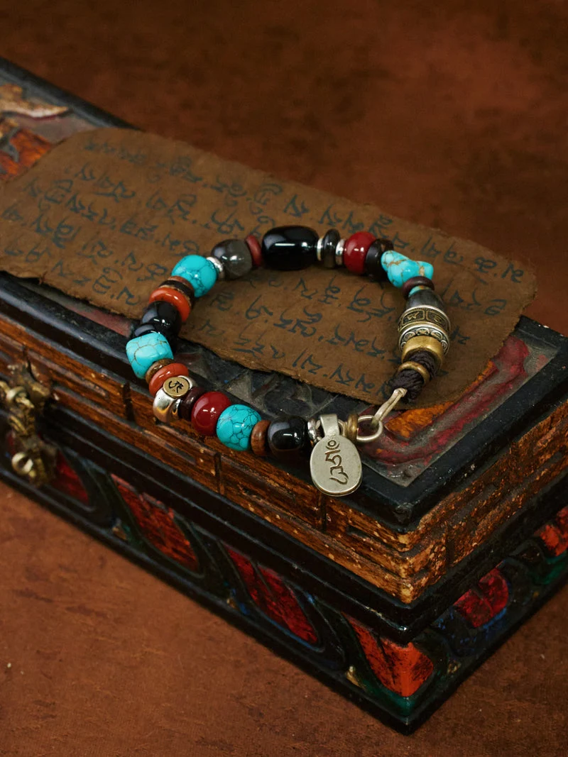 A beaded bracelet with turquoise, black, red, and metallic gold beads, featuring a silver-toned charm engraved with Tibetan script, displayed on a decorative antique-style box with a brown paper overlay.