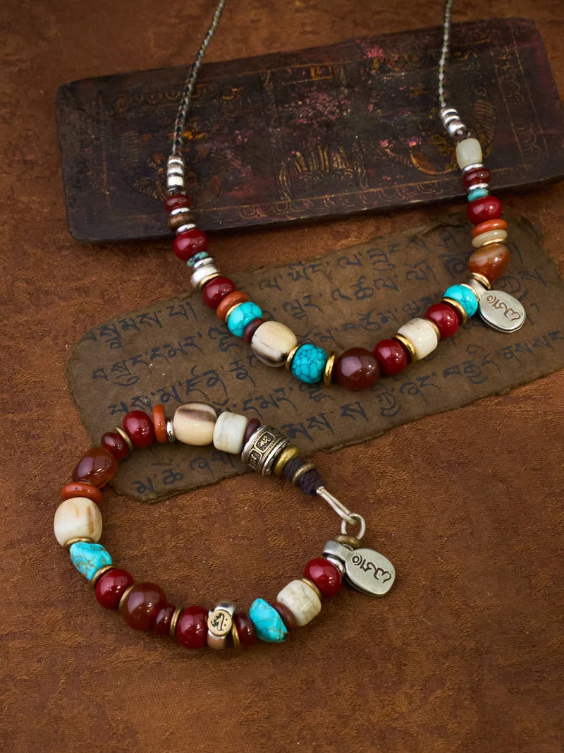 A beaded necklace and matching bracelet displayed on a textured brown surface with decorative script cards. The jewelry features dark red, turquoise blue, cream, and metallic silver beads, with a small heart-shaped silver charm engraved with Tibetan script.