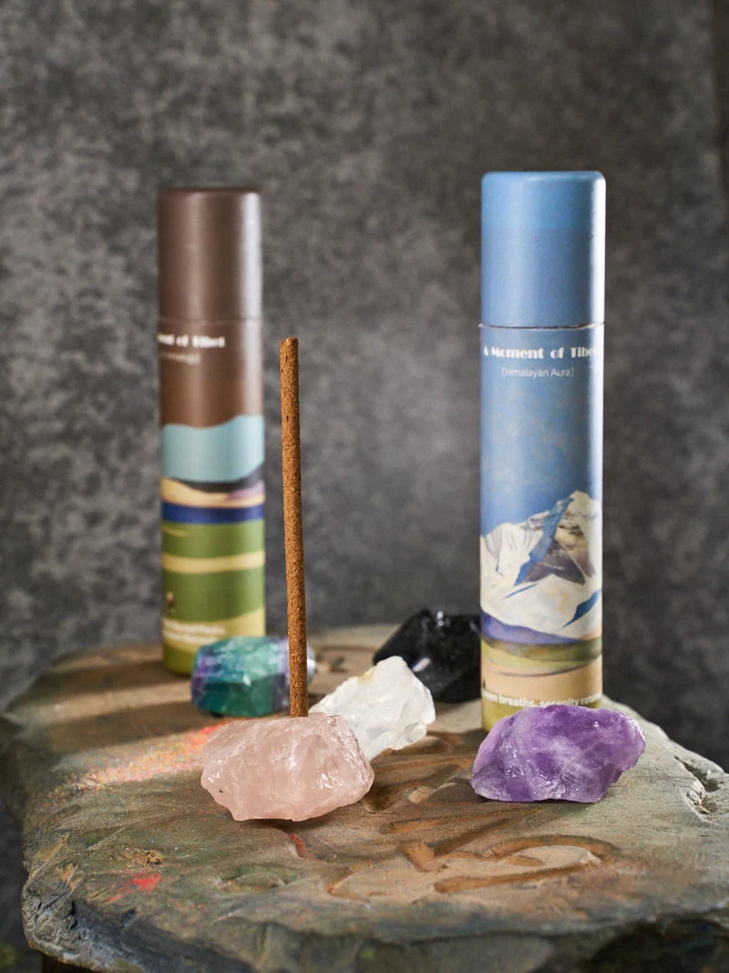 Two cylindrical incense containers with a stick and crystals on a stone surface against a dark background